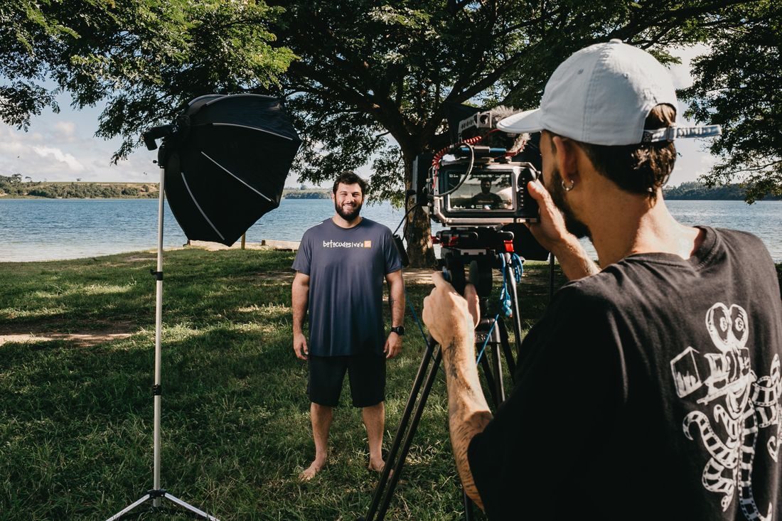 A camera operator filming a man outdoors beside a pond.