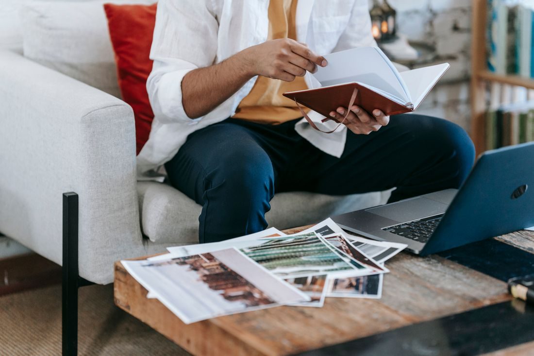 A person checking notebook notes beside photographs and a laptop.